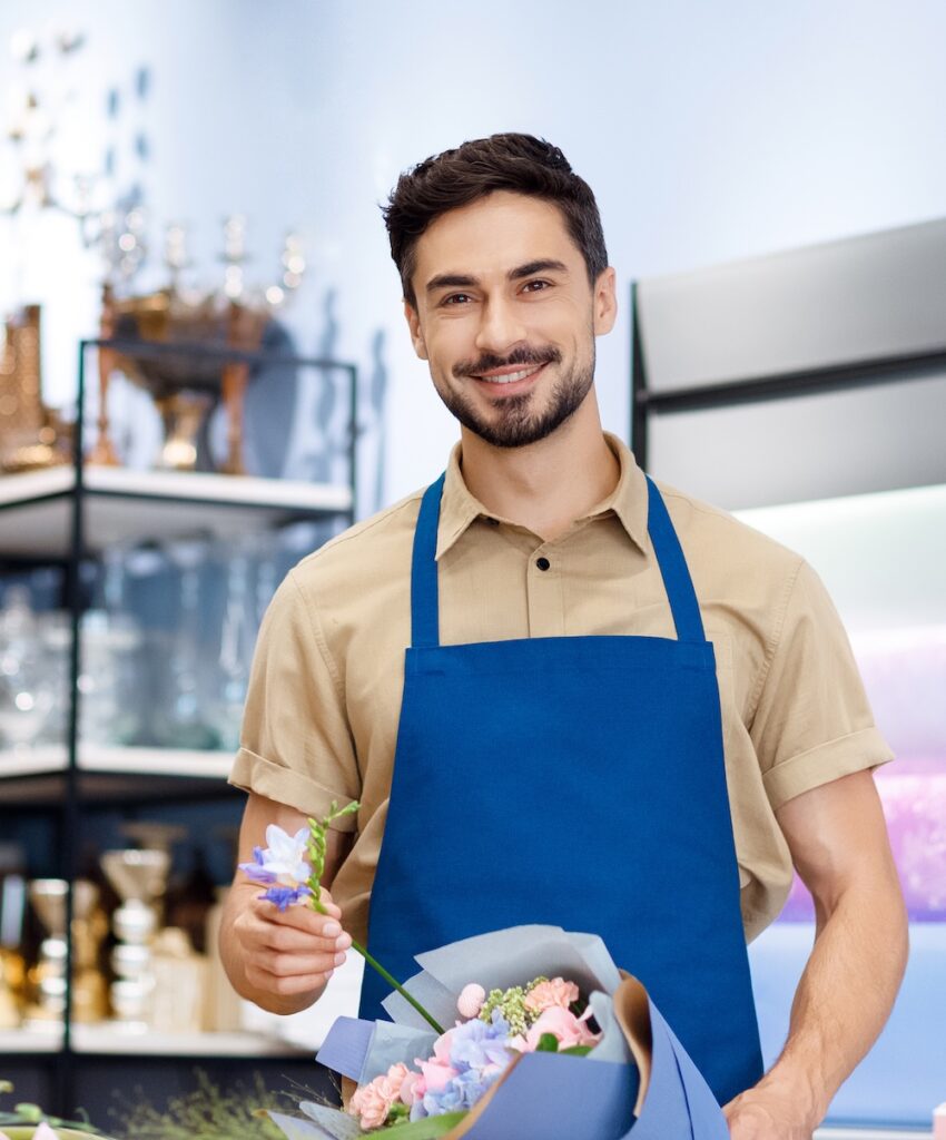 handsome bearded young florist in apron holding bouquet while working in flower shop